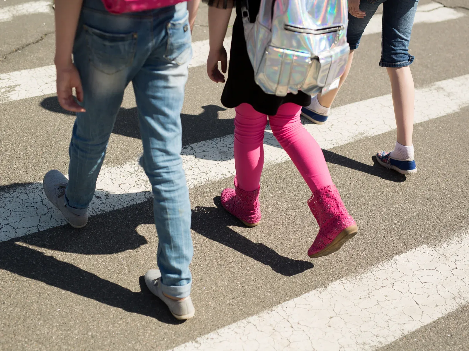 Three kids, seen from the waist down, walk across a crosswalk on a sunny day.